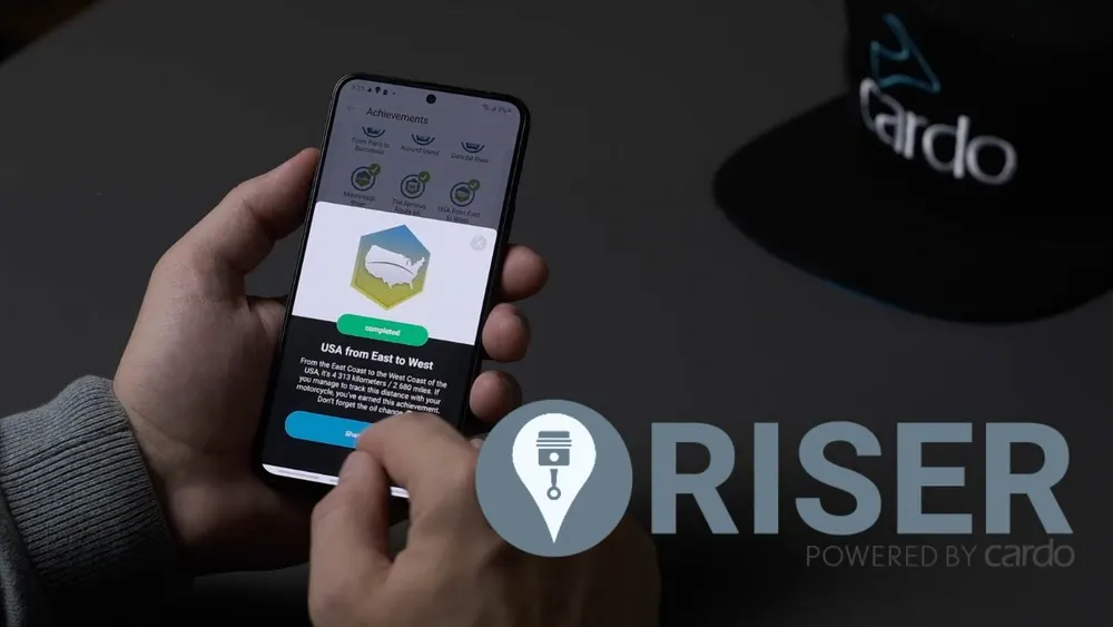 A rider holding a phone showing achievement badges in the Cardo Ride app, beside a Cardo cap
