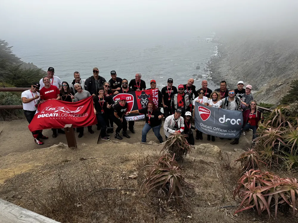 Riders gathered on a fog-covered coastal overlook at Ragged Point holding Ducati Corse and DROC flags