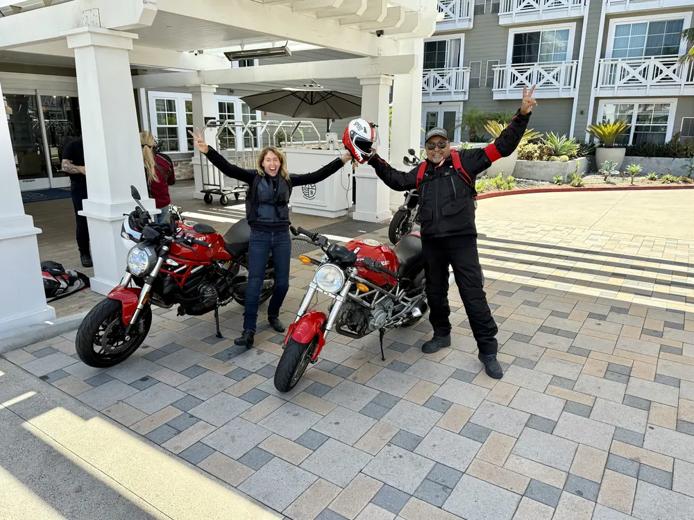Two riders with arms raised beside their Ducati Monsters at the hotel entrance in Pismo Beach
