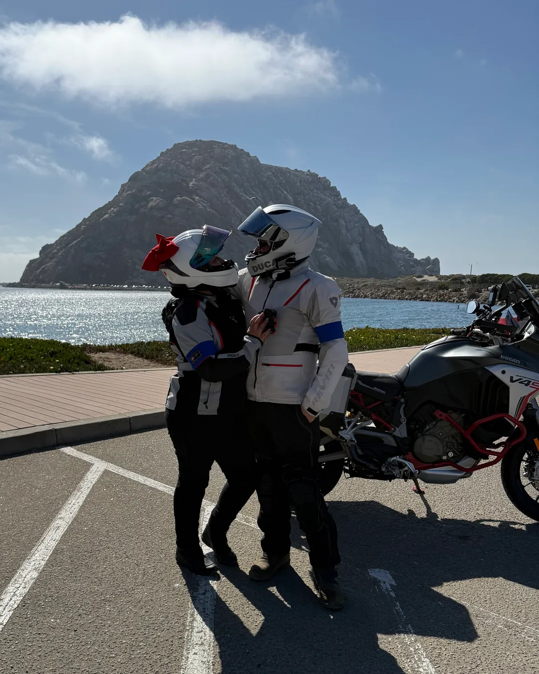 Two riders in motorcycle gear posing with their bike and Morro Rock behind them.