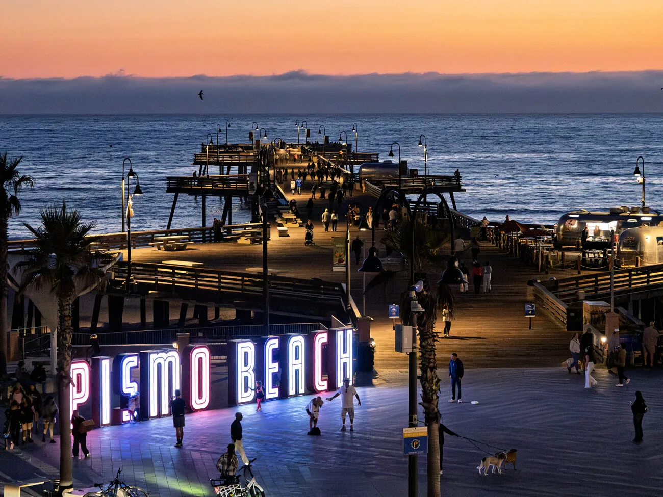 The glowing PISMO BEACH neon sign on the promenade at dusk, with the pier and ocean behind